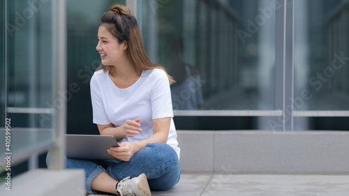 Woman in white t-shirt and blue jeans using laptop sitting on a floor outside in the city © Naypong Studio