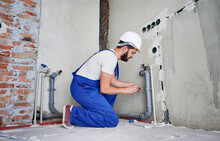 Horizontal Snapshot Of Young Plumber Working With Grey Sewer Pipes, Fixing Them To Wall With A Help Of Screwdriver. Side View Of Plumber Standing On Knees Wearing Blue Uniform And White Helmet