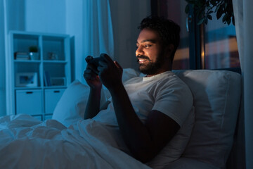 Wall Mural - technology, internet, communication and people concept - young indian man with smartphone lying in bed and playing game at home at night