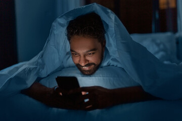 Wall Mural - technology, internet, communication and people concept - young indian man with smartphone lying in bed under blanket at home at night