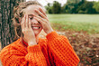 © iuricazac - Candid portrait of a gorgeous young woman smiling, hiding her face with two hands, playing peek a boo game in the park. Female wearing an orange sweater has joyful expression on the nature background.