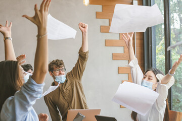 Smiling business people group,teamwork of young asian,caucasian throwing papers, documents, wearing face mask having fun, happy in office, workplace after project is done. Celebrating success concept.