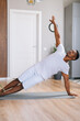 © dikushin - Fit sporty African-American man exercising in side plank position on floor during working out at bright domestic room, looking away. Concept of sport training at home gym.