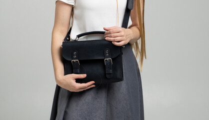 Small black leather bag over a woman shoulder on a white background. Shoulder handbag. Woman in a white shirt and grey skirt and with a black handbag. Style, retro, fashion, vintage and elegance.