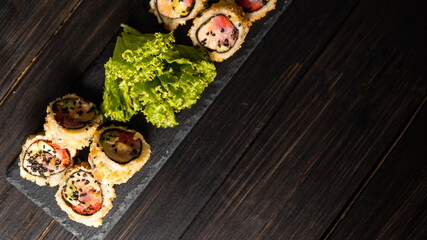 Wall Mural - Custom sushi roll in tempura with nori, fresh salmon, tuna, avocado, masago caviar, drizzled with pineapple sauce with salad pouring as decoration on a black plate on a wooden table and background.