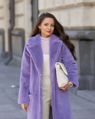 Elegant stylish woman with long brunette wavy hair wearing white trousers, pullover and shoes and purple fur coat walking city street on a sunny day