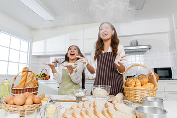 Asian mother and daughter cooking together in modern white kitchen. They throw flour into the air with funny and happy