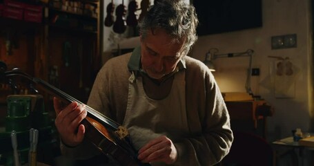 Wall Mural - Cinematic shot of experienced master artisan luthier controlling quality of handmade wood violin just made in creative workshop. Concept of spiritual instrument, art, orchestra, passion for music.