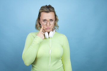 Young caucasian woman wearing headphones on neck over blue background Pointing to the eye watching you gesture, suspicious expression