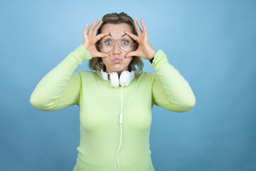 Wall Mural - Young caucasian woman wearing headphones on neck over blue background Trying to open eyes with fingers, sleepy and tired for morning fatigue