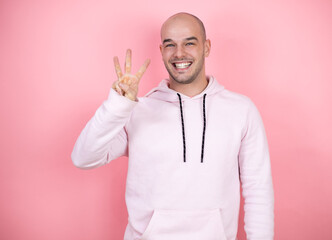 Wall Mural - Young bald man wearing casual sweatshirt over pink isolated background showing and pointing up with fingers number three while smiling confident and happy