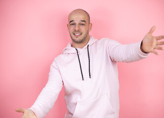 Wall Mural - Young bald man wearing casual sweatshirt over pink isolated background looking at the camera smiling with open arms for hug