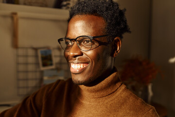 Wall Mural - Close up picture of cheerful positive young dark skinned guy in stylish eyeglasses and sweater looking with radiant smile, having optimistic facial expression, thinking about future, making plans