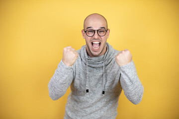 Young bald man wearing glasses over yellow background very happy and excited making winner gesture with raised arms, smiling and screaming for success.