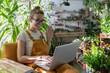 Woman gardener wear orange overalls using laptop after work, smiling and talking in video chat, say hi her friends, sitting on chair in greenhouse surrounded by exotic plants. Home gardening. 