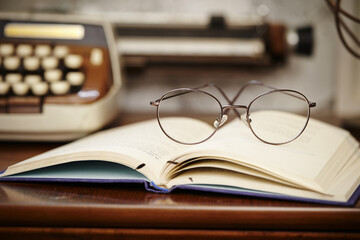 Poster - Closeup of reading glasses on an open book, with a typewriter in the background, on a wooden table