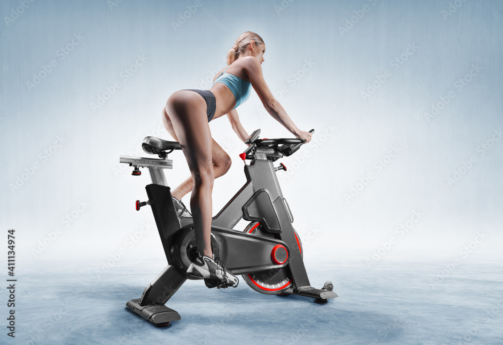 Beautiful slender and long-legged girl sits on an exercise bike. Side view. The concept of sports, bodybuilding, fitness, aerobics. healthy lifestyle. - obrazy, fototapety, plakaty 