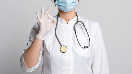 Cropped view of Woman doctor wearing protective facial mask, lab coat and stethoscope shows OK sign