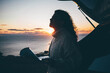Pretty curly haired woman in white sweater reads map sitting on open trunk of contemporary automobile on ocean cliff beach at bright sunset light