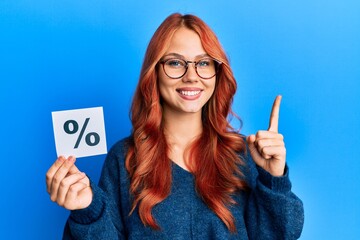 Young beautiful redhead woman holding percentage symbol smiling with an idea or question pointing finger with happy face, number one