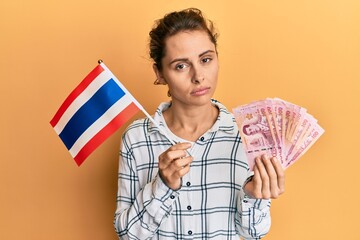 Poster - Young brunette woman holding thailand flag and baht banknotes relaxed with serious expression on face. simple and natural looking at the camera.