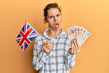 Poster - Young brunette woman holding uk flag and pounds banknotes in shock face, looking skeptical and sarcastic, surprised with open mouth