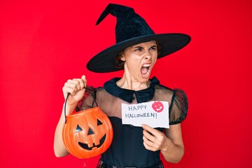 Poster - Young hispanic woman wearing witch costume holding pumpkin and happy halloween message angry and mad screaming frustrated and furious, shouting with anger. rage and aggressive concept.