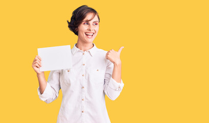 Sticker - Beautiful young woman with short hair holding blank empty banner pointing thumb up to the side smiling happy with open mouth