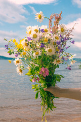 Wall Mural - a bouquet of wild flowers in your hand on the background of the river and the sky with clouds.