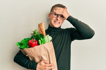 Poster - Young caucasian man holding paper bag with groceries stressed and frustrated with hand on head, surprised and angry face