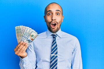 Canvas Print - Hispanic adult business man holding dollars scared and amazed with open mouth for surprise, disbelief face