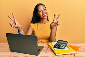 Poster - Young african american girl working at the office with laptop and calculator smiling with tongue out showing fingers of both hands doing victory sign. number two.