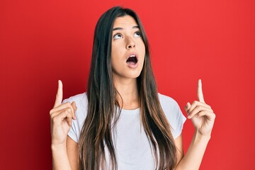 Poster - Young brunette woman wearing casual white tshirt over red background amazed and surprised looking up and pointing with fingers and raised arms.