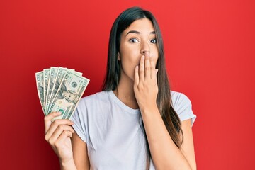 Poster - Young brunette woman holding 20 dollars banknote covering mouth with hand, shocked and afraid for mistake. surprised expression