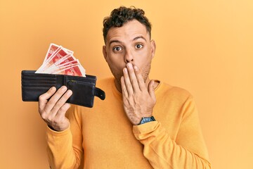 Poster - Young hispanic man holding wallet with israel shekels covering mouth with hand, shocked and afraid for mistake. surprised expression