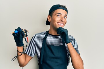 Sticker - Young handsome african american man tattoo artist wearing professional uniform and gloves holding tattooer machine with hand on chin thinking about question, pensive expression. smiling 