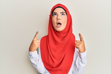 Wall Mural - Young beautiful hispanic girl wearing traditional islamic hijab scarf amazed and surprised looking up and pointing with fingers and raised arms.