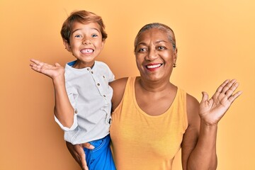 Hispanic grandson and grandmother together over yellow background celebrating achievement with happy smile and winner expression with raised hand