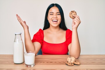 Young beautiful asian girl drinking a glass of fresh milk with chocolate cookies celebrating victory with happy smile and winner expression with raised hands