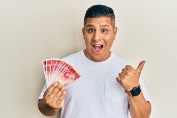 Canvas Print - Young latin man holding 20 israel shekels banknotes pointing thumb up to the side smiling happy with open mouth