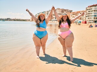 Two plus size overweight sisters twins women happy and proud of their bodies walking at the beach on summer holidays