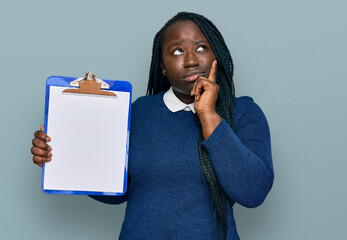 Sticker - Young black woman with braids holding clipboard with blank space serious face thinking about question with hand on chin, thoughtful about confusing idea