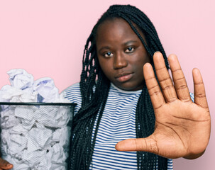 Poster - Young black woman with braids holding paper bin full of crumpled papers with open hand doing stop sign with serious and confident expression, defense gesture