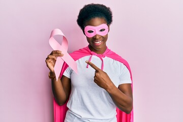 Canvas Print - Young african american girl wearing superhero costume holding pink cancer ribbon smiling happy pointing with hand and finger