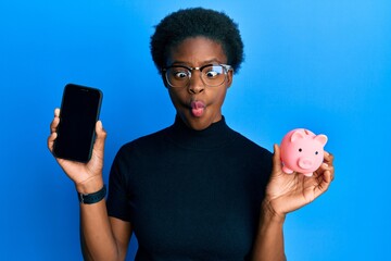 Sticker - Young african american girl holding piggy bank and smartphone making fish face with mouth and squinting eyes, crazy and comical.