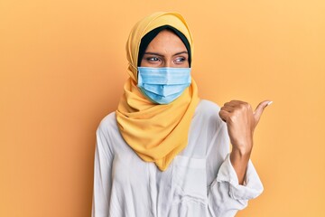 Canvas Print - Young brunette arab woman wearing traditional islamic hijab and medical mask smiling with happy face looking and pointing to the side with thumb up.