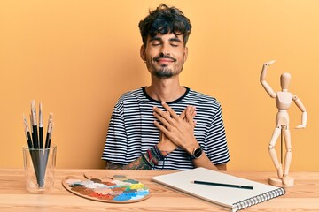 Sticker - Young hispanic man sitting on the table at art studio smiling with hands on chest with closed eyes and grateful gesture on face. health concept.