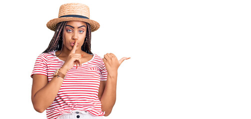 Poster - Young african american woman with braids wearing summer hat asking to be quiet with finger on lips pointing with hand to the side. silence and secret concept.