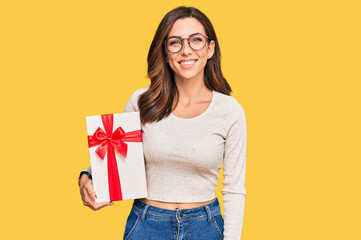 Young brunette woman holding gift looking positive and happy standing and smiling with a confident smile showing teeth