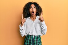 Beautiful African American Woman With Afro Hair Wearing Scholar Skirt Amazed And Surprised Looking Up And Pointing With Fingers And Raised Arms.